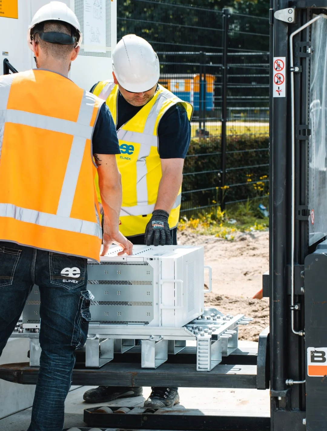 Twee bouwvakkers met veiligheidshelmen en reflecterende vesten verplaatsen een groot wit apparaat met een vorkheftruck op een bouwplaats.